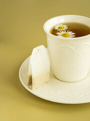 Сhamomile tea in a white cup. Tea bag on a white saucer. Сhamomile flowers on tea surface. Yellow...