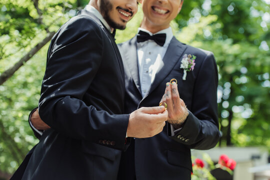 partial view of happy gay couple in formal wear holding golden wedding rings in hands.