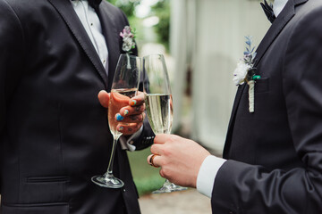 cropped view of gay newlyweds in suits holding glasses with champagne on wedding day.