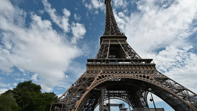 Paris, France. June 2022. Amazing wide angle lens footage at the base of the Eiffel tower. Beautiful summer day with blue sky and white clouds. The imposing base of the tower dominates the scene.