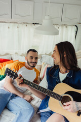 happy and tattooed gay man playing acoustic guitar near boyfriend and lgbt flag on bed in van.