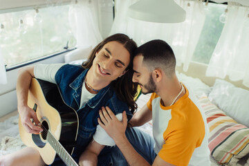 high angle view of happy gay man with long hair playing acoustic guitar near boyfriend on bed in modern van.