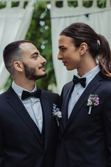 side view of gay couple in suits with boutonnieres looking at each other during wedding ceremony.