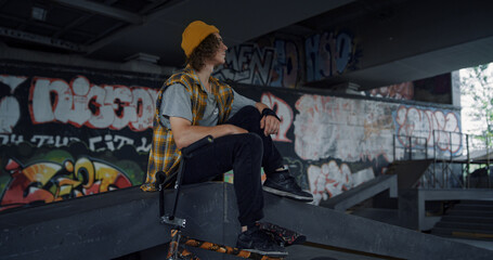 Calm teenager relaxing on ramp at skatepark. Handsome man having break.