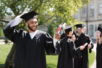 Cheerful african american bachelor looking at diploma near blurred friends in park.