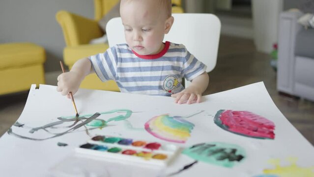 baby child boy kid learning to paint using paintbrush and aquarelle, early home education Montessori lessons for toddlers. caucasian baby child sitting in living room drawing artwork