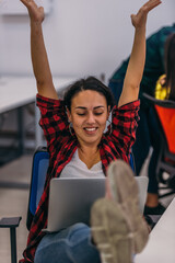 A young female worker with her hands up in the air, her legs on the office desk and her laptop on her lap, is excited for launching her project.
