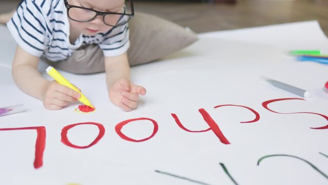 close-up child boy in glasses drawing poster back to school words lying on floor in room. cute caucasian elementary preschooler getting ready for first day in school, nursery, kindergarten education