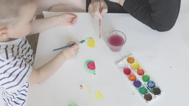 happy family home education concept mother teaching son to paint draw watermelon, using paintbrush and water aquarelle paints. caucasian child kid boy learning drawing, painting fruits together mom