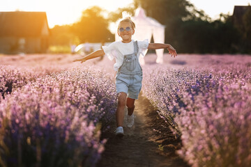 happy child girl is walking in lavender field on sunset. Smiling kid in suglasses , jeans jumpsuit is having fun in nature on summer day. Cheerful little girl. happy childhood concept