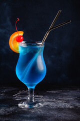 Glass of blue lagoon cocktail decorated with orange and cherry at festive bar counter background.