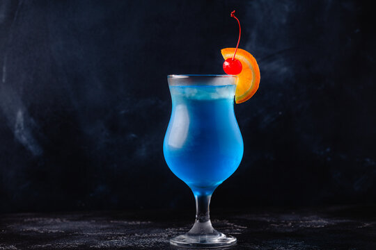 gGass of blue lagoon cocktail decorated with orange and cherry at festive bar counter background.