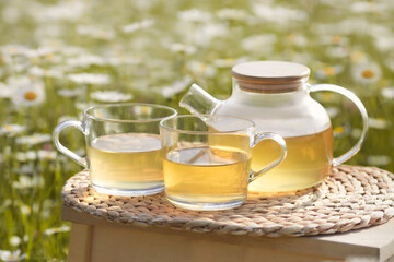 Chamomile herbal tea in transparent teapot and two glass cup on table on flowers field background
