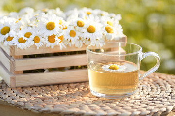 Obraz na płótnie Canvas Glass cup of chamomile hot tea on background with wooden crate full of flowers outdoors
