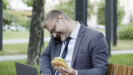 Multitasking male freelancer talking on phone, working on laptop, eating burger