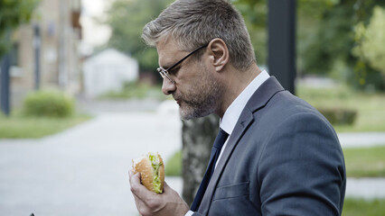 Hungry office worker eating burger in park during a break, unhealthy lifestyle