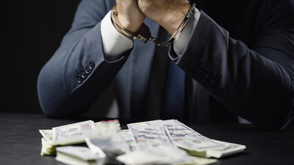 Businessman in elegant suit sitting handcuffed, lots of money on desk, financial fraud