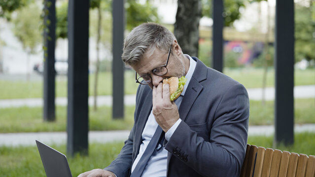 Sales manager working on laptop, sitting on bench in park and eating a burger