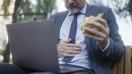 Male manager feeling stomachache after eating burger on lunch in park, indigestion