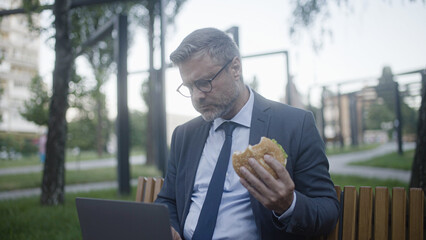Man in suit eating burger while working on laptop outdoor, unhealthy food habits