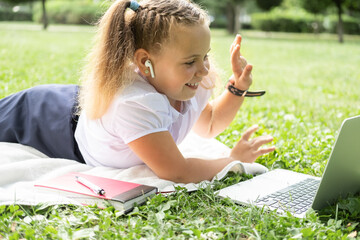 happy kid girl on school uniform with wireless headphone has online lesson on laptop on lawn