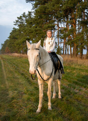 Little smiling girl riding  white blue eyed horse in the autumn evening forest 