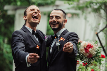 excited gay couple in formal wear holding sparklers on wedding day.