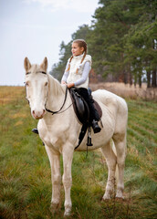 Fototapeta premium Cute girl sitting on a white blue eyed horse in the autumn forest 