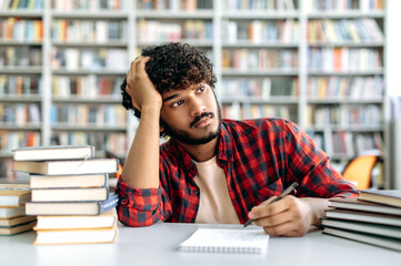 Sad arabian or indian male student tired from studying, sits at a desk in a library against the...