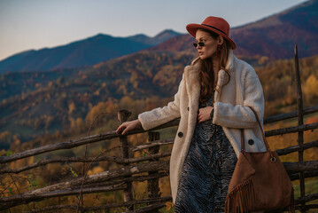 Fashionable woman wearing trendy autumn outfit with orange hat, glasses, faux fur coat, holding trendy brown suede bag, posing in mountain  landscape. Copy, empty space for text