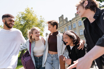Smiling student hugging interracial friends with backpacks outdoors.