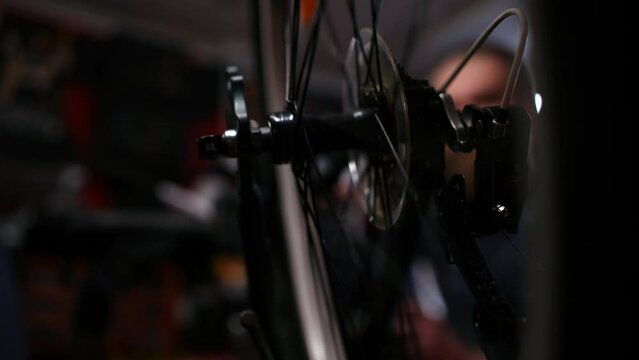Close-up tracking shot of bicycle repairman adjusting levers on handlebar and inspecting spinning rear wheel an inverted bike, working in dark garage of professional repair shop, slow motion.