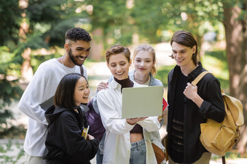 Smiling student holding laptop near interracial friends in summer park.