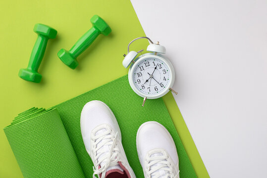 Fitness concept. Top view photo of white sneakers over green exercise mat dumbbells and alarm clock on bicolor green and white background with copyspace