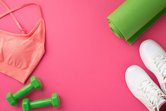 Sports accessories concept. Top view photo of green exercise mat pink sports top dumbbells and white shoes on isolated pink background with copyspace
