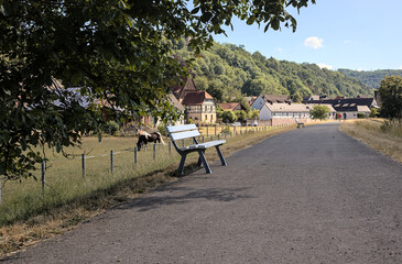 White bench under a tree on a small road in a rural setting with half-timbered houses and horses and mountains with forest and trees and two people at a great distance hiking