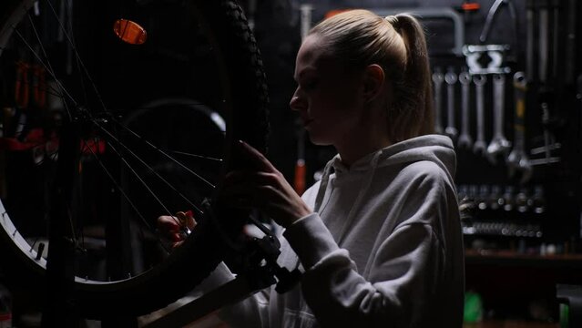 Portrait of young pretty woman aligning tension spokes of bicycle wheel with special tool in dark garage of professional repair shop. Skilled serviceman performing diagnostics of MTB, slow motion.