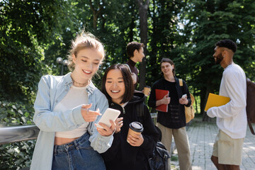 Smiling student pointing with finger at smartphone near asian friend with coffee to go in park.
