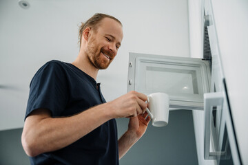 Young ginger man using microwave while cooking food in kitchen