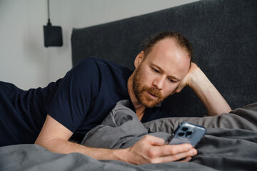 Young ginger man using mobile phone while resting on bed