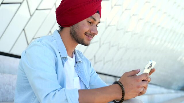 Portrait of cheerful young Indian guy in turban sending message on mobile phone sitting on the steps outdoors. Happy ethnic man using smartphone, enjoying chatting online, messaging in social networks