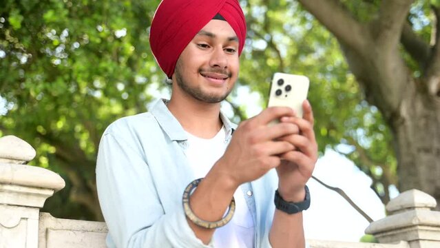 Carefree young Indian guy in casual shirt and traditional headdress using smartphone outdoors. Male student texting with classmates online, scrolling news feed in social networks, checking schedule