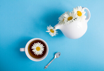 Top view of white tea kettle with chamomile and tea cup on the blue background. Closeup. Copy space.