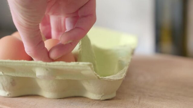 Person taking eggs from an egg box. The box is light green and the eggs are brown. The egg box is on a wooden cutting board.