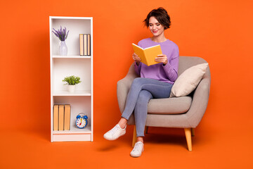Full length photo of shiny funny girl dressed purple pullover reading story sitting armchair isolated orange color background