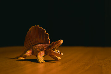 A figurine of an aggressive growling orange dinosaur of the Dimetrodon species stands on a brown table against a dark background