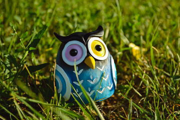 A toy of a surprised blue-colored owl with multi-colored eyes close-up stands in the middle of green grass in the light of the evening sun