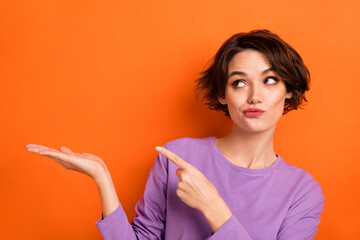 Photo of doubtful unsure lady wear violet sweatshirt holding arm pointing looking empty space isolated orange color background