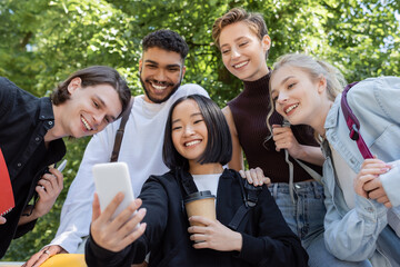 Asian student holding coffee to go and taking selfie with interracial friends in park.