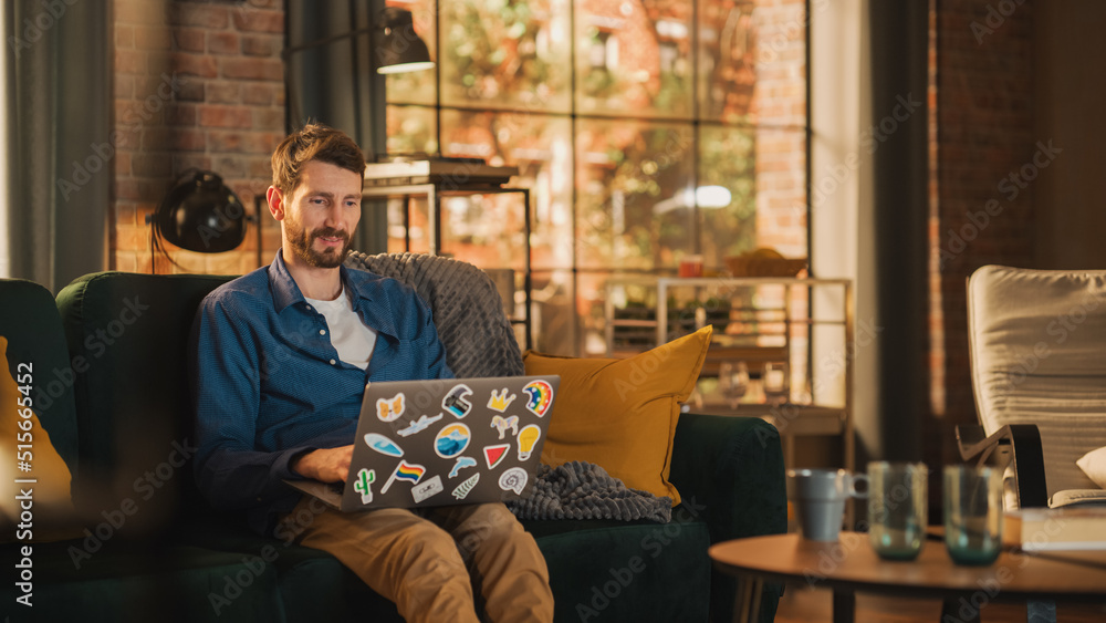 Poster portrait of a handsome man sitting on couch, using laptop computer in stylish loft apartment on a su